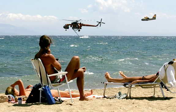 Туристы загорают в то время, как вертолет набирает воду для тушения пожаров в Греции