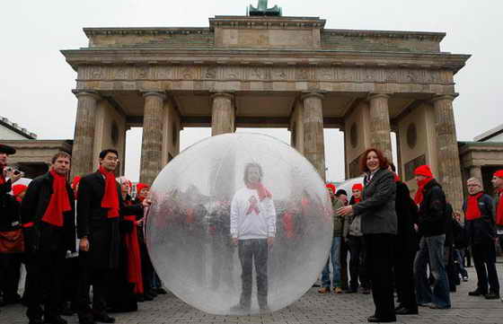 Учстник кампании по борьбе со СПИДом стоит в пластиковом пузыре в окружении работников здравоохранения перед Бранденбургскими воротами в Берлине. Пузырь символизировает стигматизацию людей, живущих с ВИЧ и СПИДом
